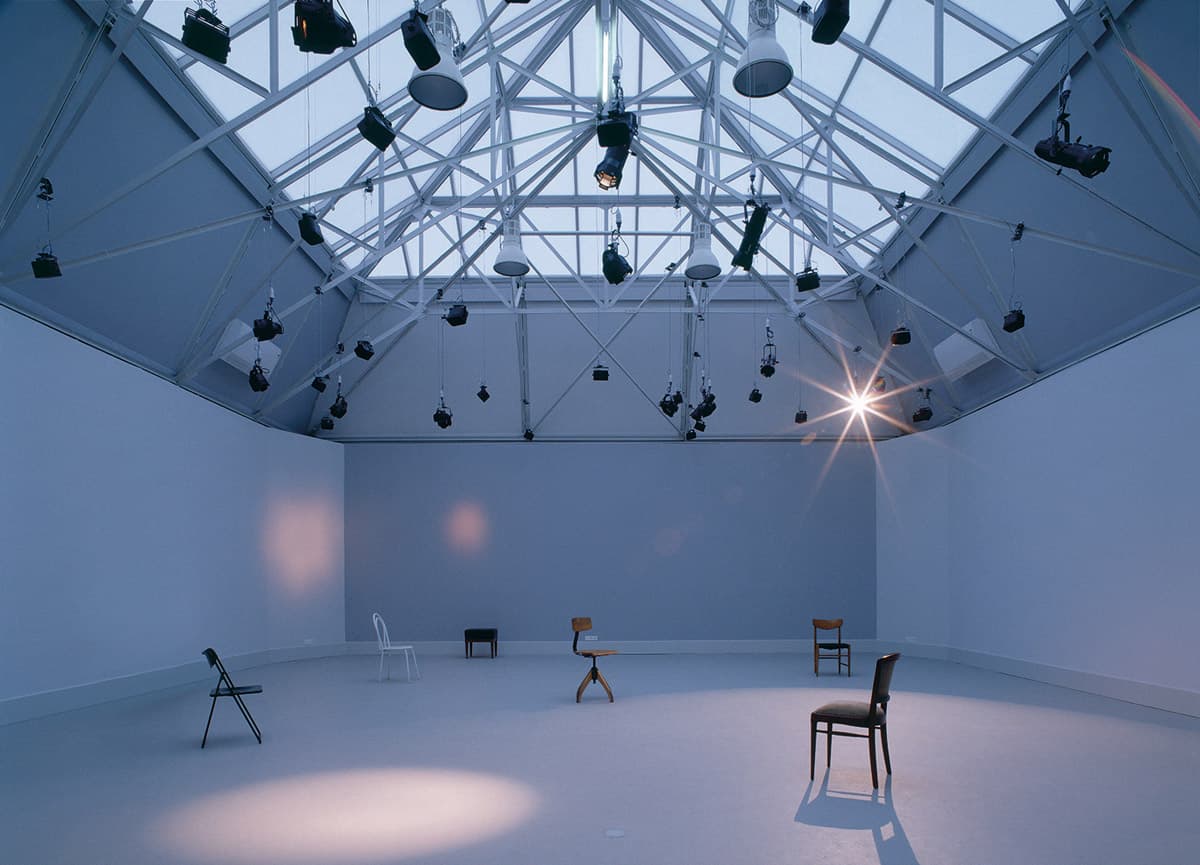 Ausstellungsraum im Dämmerlicht mit leeren Wänden und über die Fläche verteilten Stühlen, an der Lichdecke hängen Scheinwerfer