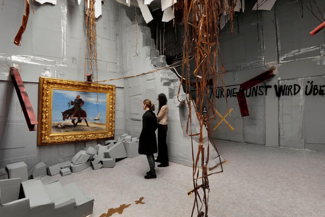 Ruinenartiger Ausstellungsraum, vorne hängen kupferfarbene Bänder von der Decke, im Hintergrund stehen zwei Frauen und betrachten ein Gemälde. 