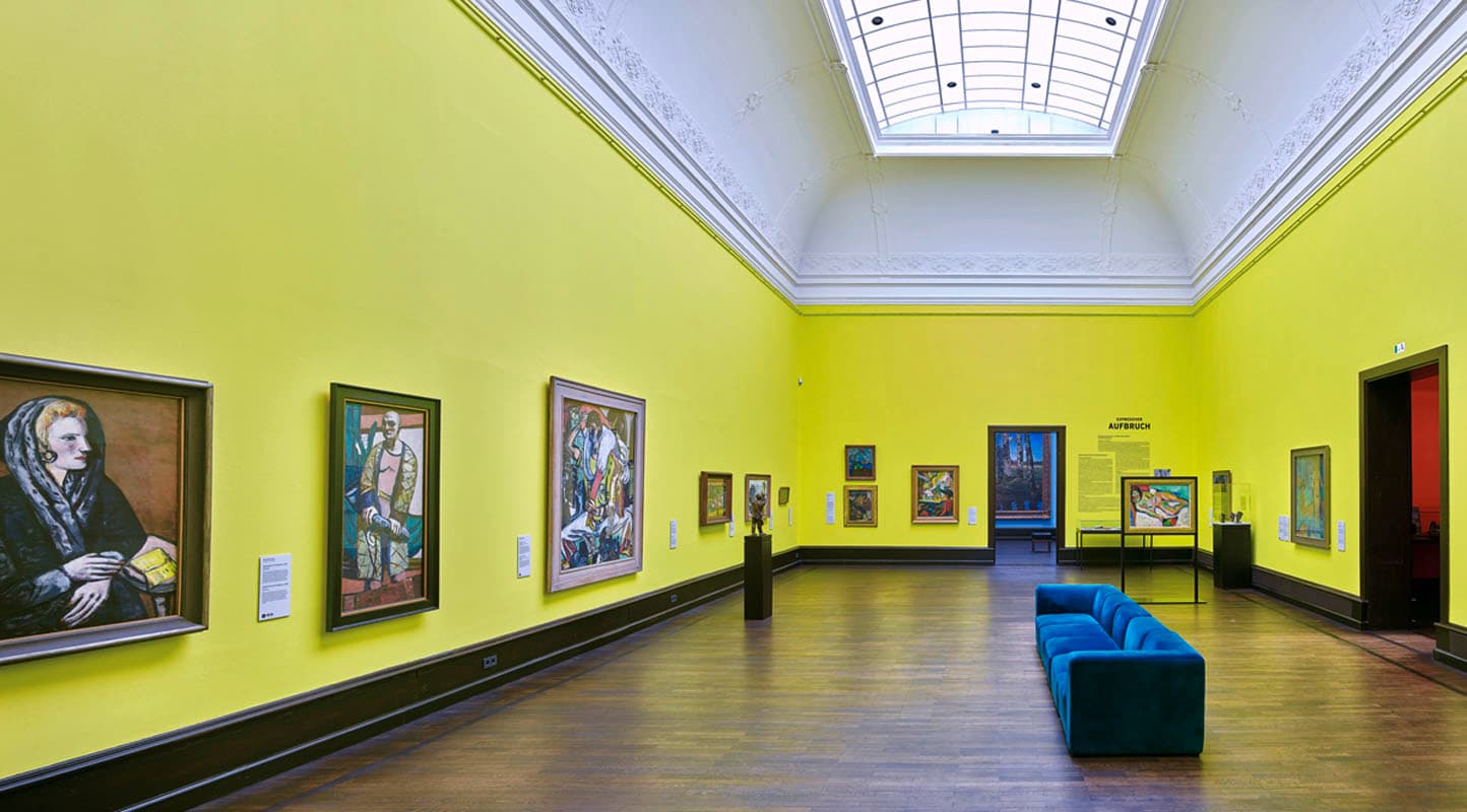 Ausstellungsraum mit bunten Gemälden an zitronengelben Wänden und einem blauen Sofa in der Mitte