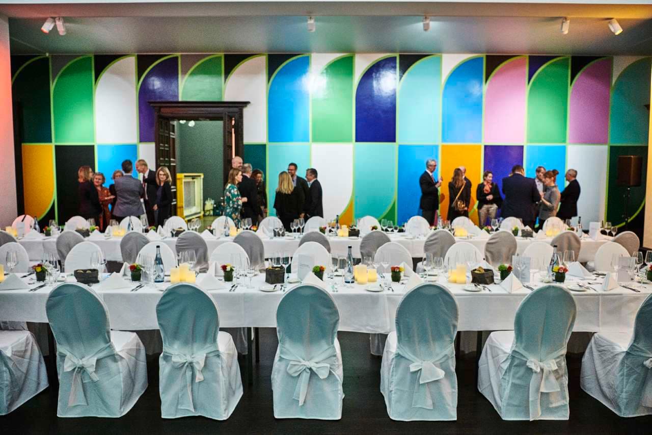 Raum mit festlich gedeckten langen Tischen, im Hintergrund unterhalten sich Personen vor einer Wand mit buntem grafischen Muster.