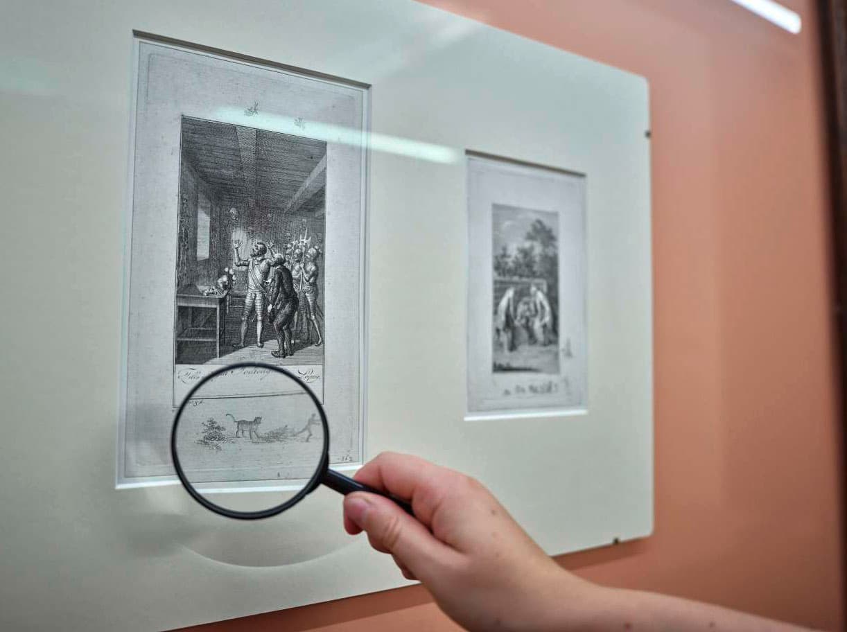 Eine Hand hält eine Lupe, die ein Detail am Rande einer schwarz-weißen Grafik in einem Passepartout vergrößert.