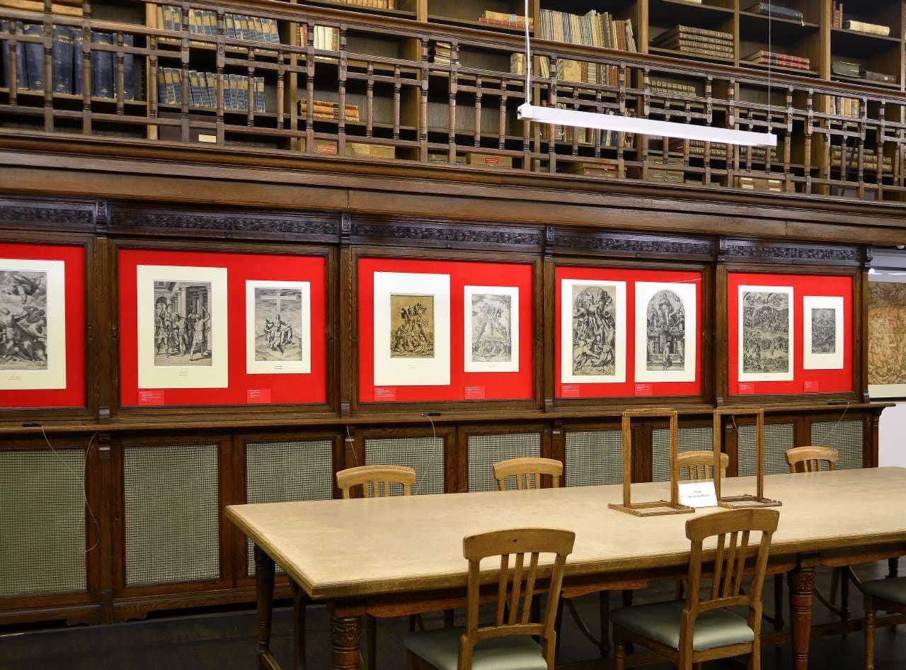 Raumansicht mit Tisch und Holzstühlen, im Hintergrund Werke auf Papier auf rotem Untergrund, darüber eine Holzgalerie mit Büchern