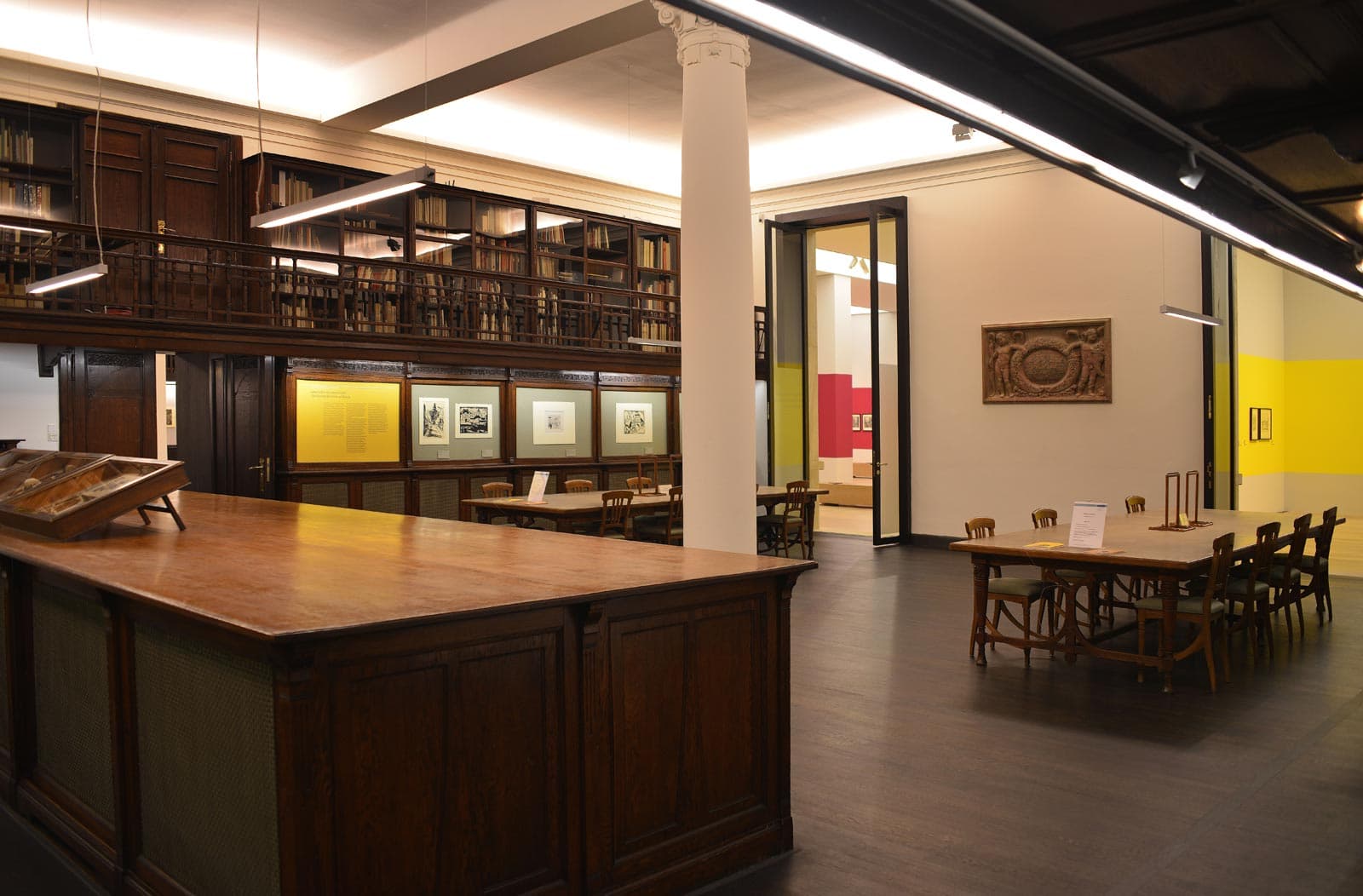 Eine Raum mit dunklen Holztischen, Glasvitrinen an der linken Wand und einer weißen Säule in der Mitte