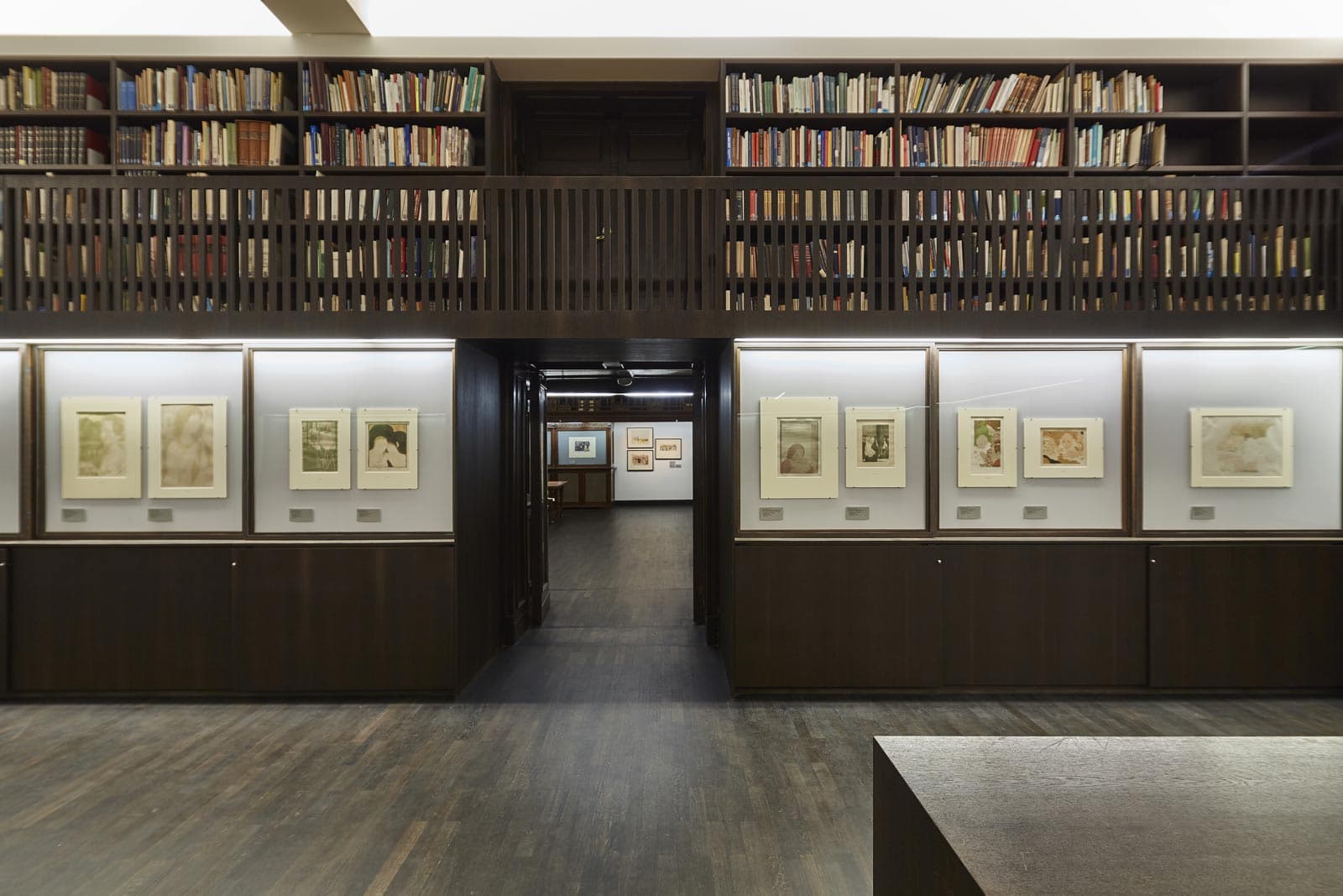 Ein Raum mit dunklem Holzboden,Glasvitrinen an der Wand, in denen Kunst in Passepartouts hängt, darüber dunkle Holzregale mit Büchern.