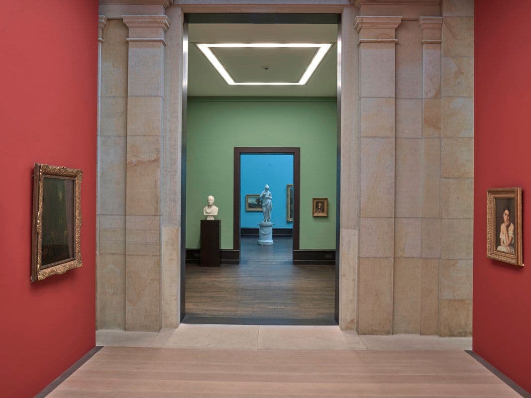 Blick durch mehrere Galerieräume, vorne rote Wände, dahinter grüne und ganz im Hintergrund eine Skulptur vor hellblauer Wand