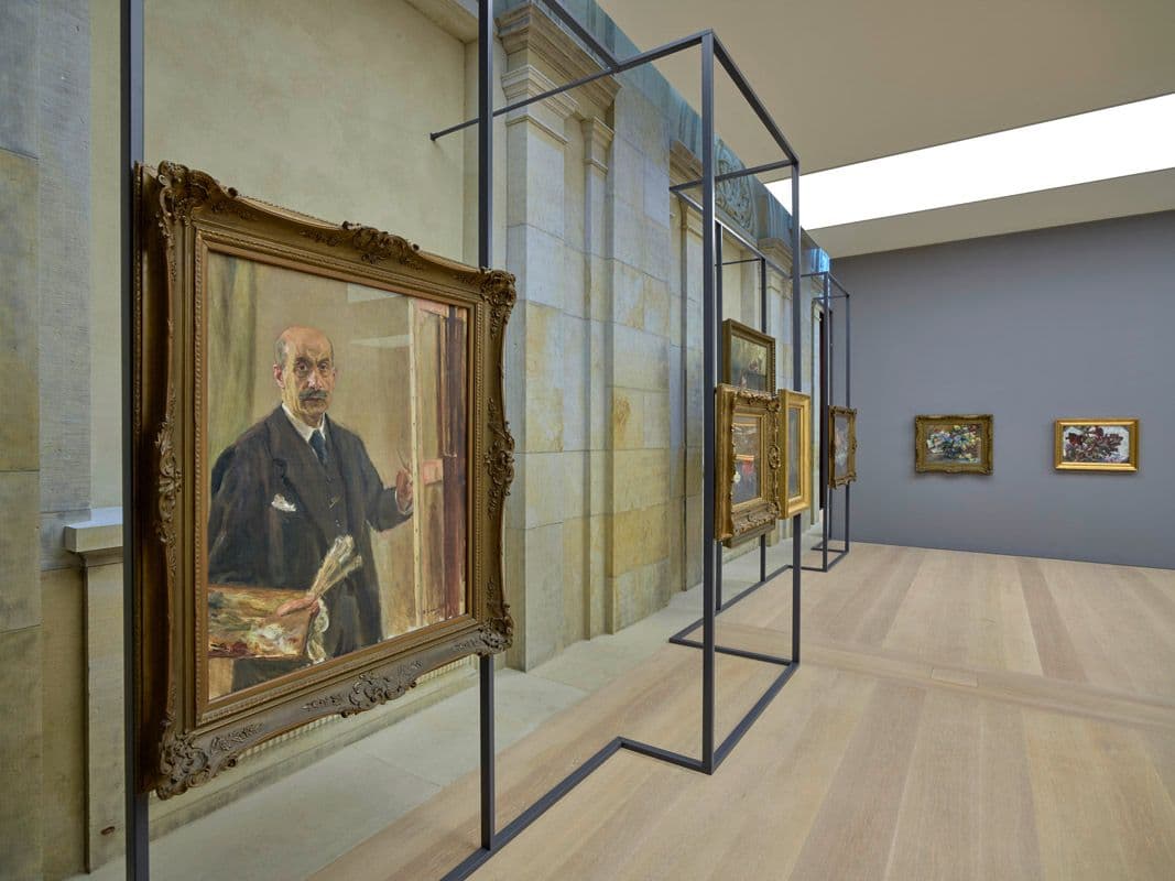 Ausstellungsraum mit Gemälden an einem scharzen Gerüst an Sandsteinwand