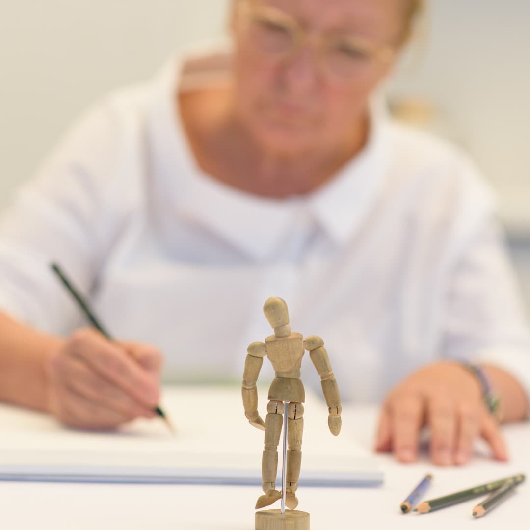 Eine Frau zeichnet, im Vordergrund steht eine kleine Holzfigur.